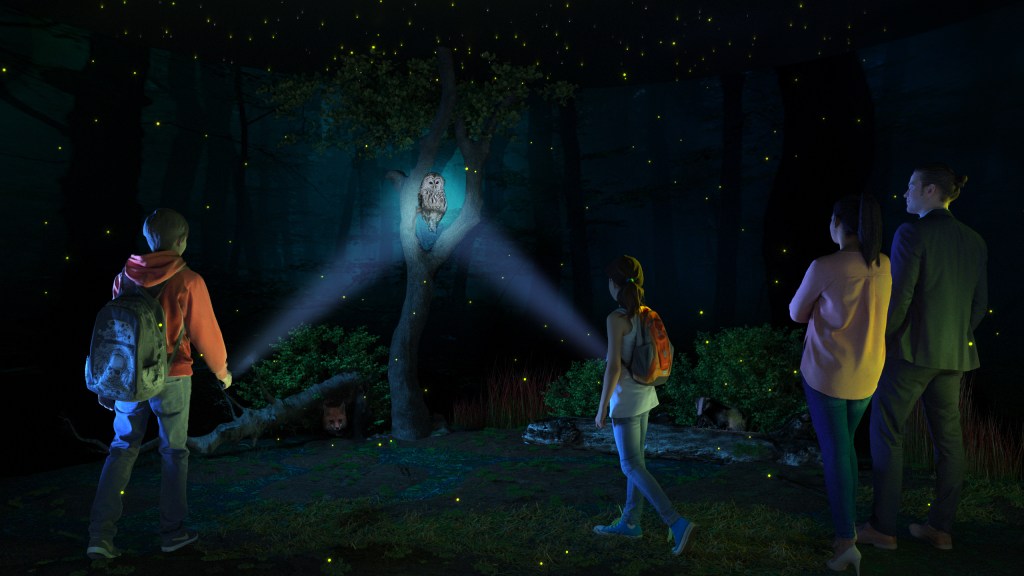 A group of people walking in a dark exhibition room that ressembles a natural envirnoment. Two children point their torches at an owl perched on a tree while the adults look from the side.